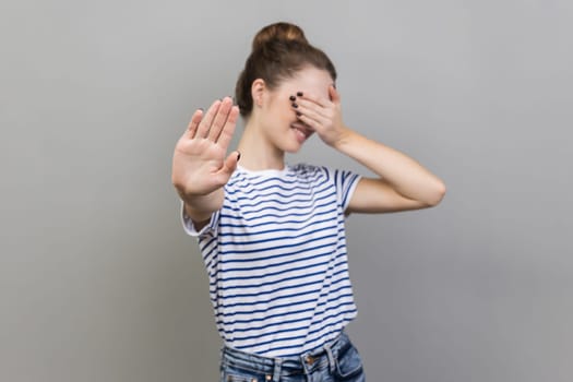 Portrait of afraid beautiful woman wearing striped T-shirt closing eyes with palm and showing stop hand gesture, turning face, does not want to see. Indoor studio shot isolated on gray background.