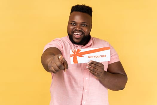 Portrait of excited amazed bearded man wearing pink shirt holding gift vaucher pointing to camera with toothy smile, you win present. Indoor studio shot isolated on yellow background.