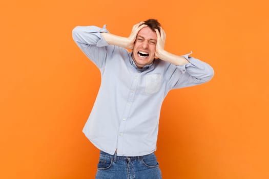 Portrait of upset sad young adult man standing with hands on head, suffering terrible headache, screaming from pain, wearing light blue shirt. Indoor studio shot isolated on orange background.