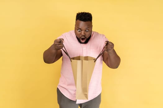 Portrait of man wearing pink shirt holding looking inside shopping bag, being astonished with cheap purchase or excellent gift. Indoor studio shot isolated on yellow background.