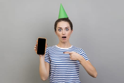 Portrait of surprised astonished woman wearing striped T-shirt and party cone, showing pointing mobile phone with empty black screen for advertisement. Indoor studio shot isolated on gray background.