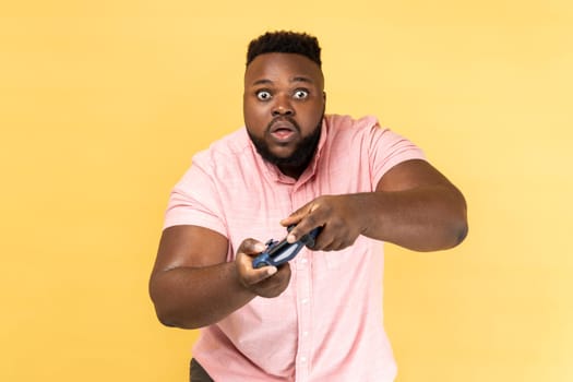 Portrait of shocked surprised bearded man wearing pink shirt playing games, holding joypad, looking at camera with big eyes and open mouth. Indoor studio shot isolated on yellow background.