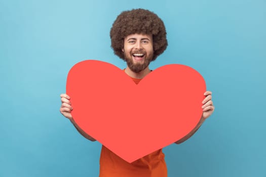 Portrait of man with Afro hairstyle wearing orange T-shirt holding big red heart, expressing positive romantic emotions, looking at camera with smile. Indoor studio shot isolated on blue background.