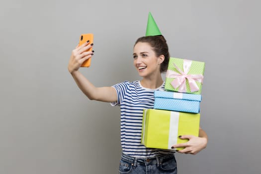 Portrait of friendly smiling woman wearing striped T-shirt and party cone, holding stack of present boxes and taking selfie, broadcasting livestream. Indoor studio shot isolated on gray background.