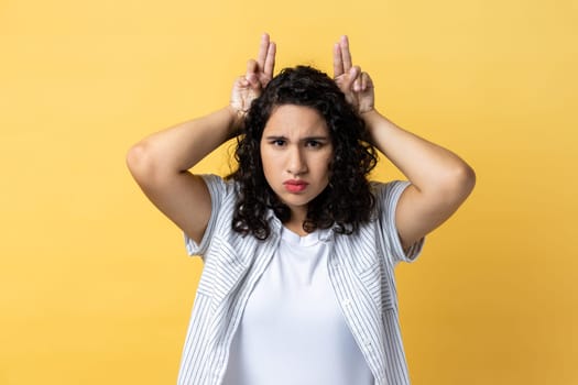 Portrait of strict serious woman with dark wavy hair showing bull horn gesture with fingers over head, looking hostile and threatening. Indoor studio shot isolated on yellow background.