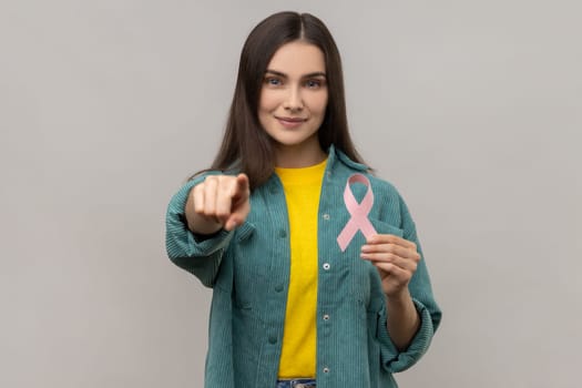 Portrait woman holding pink ribbon, symbol of breast cancer awareness, looking and pointing at camera, wearing casual style jacket. Indoor studio shot isolated on gray background.