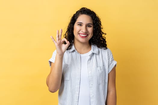 Portrait of smiling satisfied positive optimistic beautiful woman with dark wavy hair looking at camera showing ok sign gesture. Indoor studio shot isolated on yellow background.