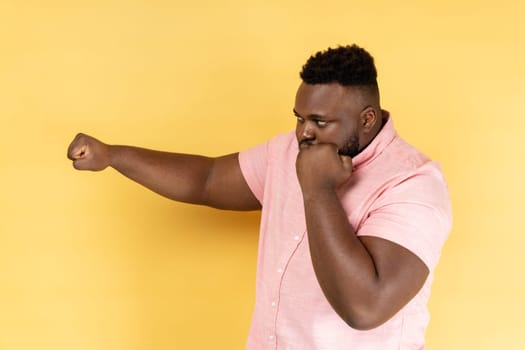 I'll hit you. Side view of bearded man wearing pink shirt standing with raised fists boxing gesture ready to punch, ready to struggle, fighting spirit. Indoor studio shot isolated on yellow background