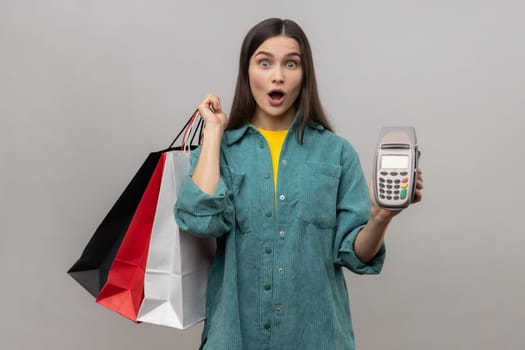 Shocked surprised woman with dark hair holding payment terminal and paper shopping bags, easy express order and delivery, wearing casual style jacket. Indoor studio shot isolated on gray background.