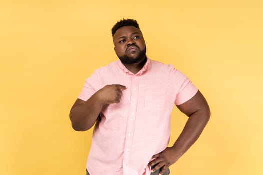 Portrait of young adult bearded man wearing pink shirt pointing fingers on himself and looking away with pride, extremely confident. Indoor studio shot isolated on yellow background.