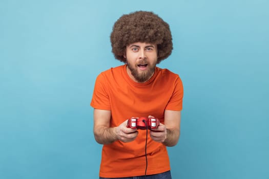 Portrait of concentrated excited man with Afro hairstyle grimacing holding joystick, playing video games online, virtual competition. Indoor studio shot isolated on blue background.