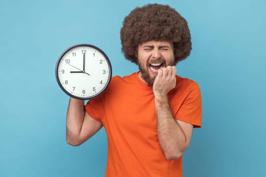 Portrait of man with Afro hairstyle wearing orange T-shirt biting nails on his fingers holding big wall clock in hand, worried about deadline. Indoor studio shot isolated on blue background.