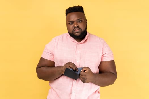 Portrait of sad poor depressed bearded man wearing pink shirt standing looking at camera with upset facial expression, showing empty wallet. Indoor studio shot isolated on yellow background.