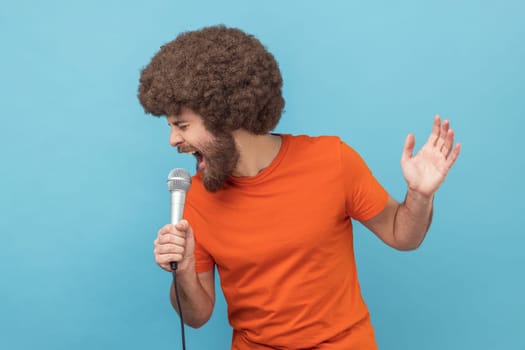 Portrait of man with Afro hairstyle wearing orange T-shirt singing songs, holding microphone, singer making performance, having fun on party in karaoke. Indoor studio shot isolated on blue background.