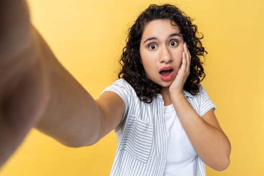 Portrait of shocked surprised woman with dark wavy hair making point of view photo, looking at camera with open mouth, keeps hand on cheek, POV. Indoor studio shot isolated on yellow background.