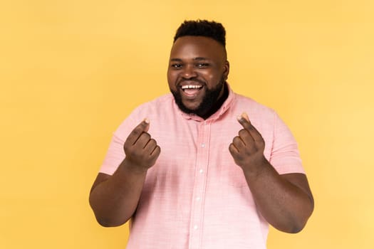 Portrait of smiling bearded man wearing pink shirt makes money gesture, rubs fingers, looking at camera with glad expression. Indoor studio shot isolated on yellow background.