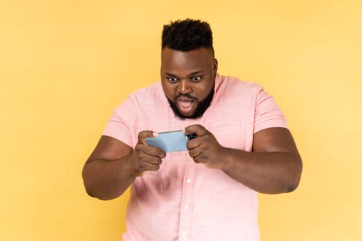 Portrait of amazed man wearing pink shirt standing, using smartphone and playing mobile game with excited face and big eyes. Indoor studio shot isolated on yellow background.