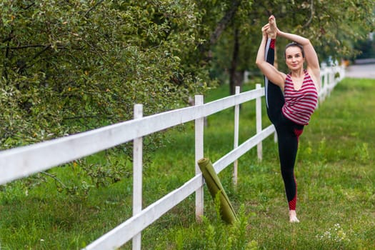 Full length portrait of flexible young woman wearing sportswear, stretching her leg, balancing, practicing yoga, having workout outdoor, raising leg in air, doing split stretch one foot.