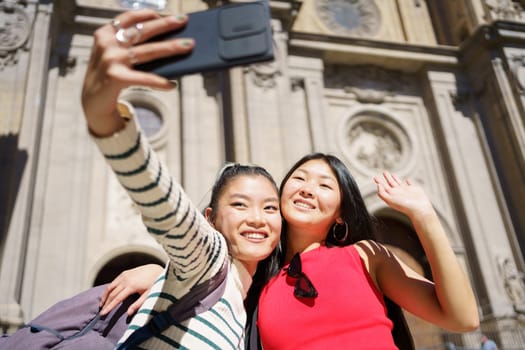 Happy young Asian female travelers, in casual clothes with backpacks smiling and hugging while waving hands and taking selfie on smartphone during summer vacation in Spain