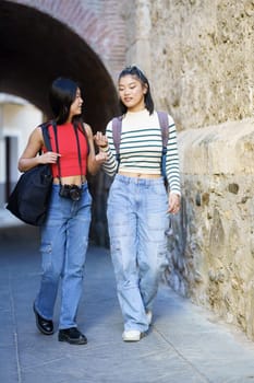 Full body of young Asian female tourists in casual clothes with bags and photo camera strolling against stone wall while talking to each other