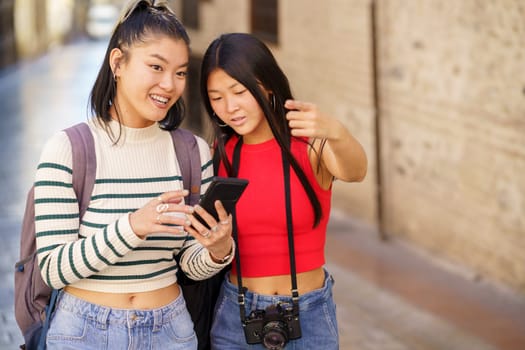 Asian girls in their 20s traveling through city of Granada using map on cellphone and pointing away while looking for direction during trip together