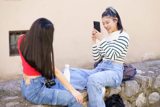 Positive young ethnic woman in striped sweater and jeans, sitting on stone border against girlfriend with long black hair and taking photo on smartphone during trip