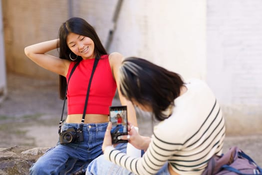 Positive young ethnic woman in red top and jeans touching hair while posing for photo in front of girlfriend in striped sweater during sightseeing