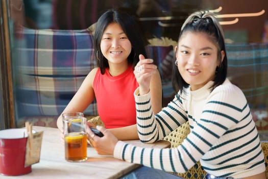 Positive young Asian female friends in casual clothing, smiling and looking at camera while chilling together at table with refreshing drinks and sharing smartphone in cafe