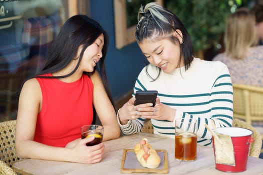 Content young Asian female friends in casual outfits, smiling and taking photo of delicious meal on smartphone while gathering at wooden table with glasses of drinks in cafe