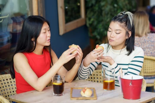 Calm young Asian female friends in casual apparel having lunch time, in cafe together while eating yummy food and drinking refreshing beverages during meeting with each other