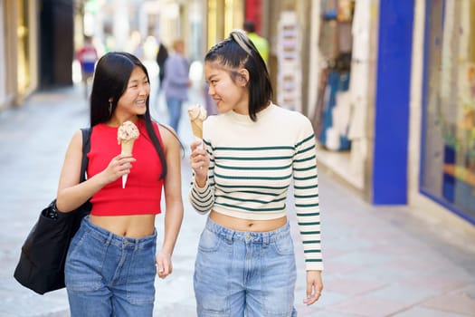 Happy young Asian girlfriends smiling and eating delicious ice cream cone while walking together on street in city
