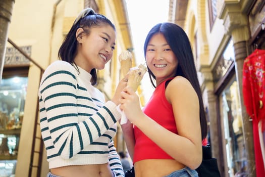 Low angle of positive Asian female friends eating tasty ice cream while standing on street and enjoying time together in daytime