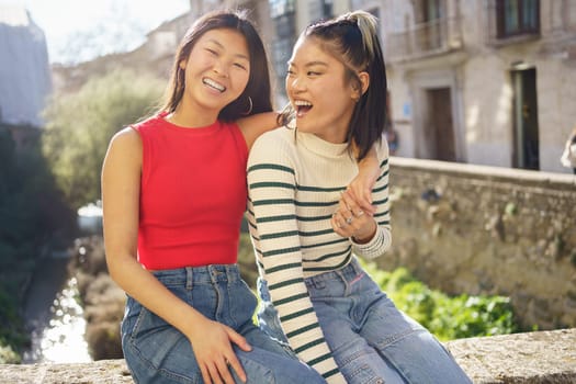 Happy young Asian female putting hand on shoulder of friend in casual clothes smiling and looking at camera while sitting on stone bench in Granada