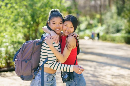 Cheerful young Asian female tourists in casual clothes with backpack and camera smiling, while hugging and looking at camera during trip in sunny forest of Alhambra