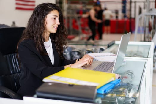 Glad female with dark wavy hair smiling and using laptop while sitting at desk during psychology session in gym