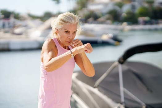 Mature woman doing sport in a coastal port. Arm training throwing boxing punches. Female doing shadow boxing outdoors.