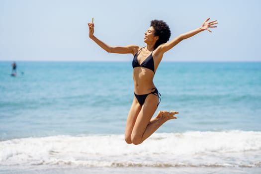 Full body side view of cheerful African American female traveler in bikini taking self portrait on smartphone and jumping on coast near waving sea