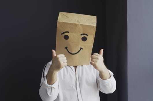 A man in a white shirt with a paper bag on his head, with a smiley face drawn, stands against the wall, overwhelmed with feelings, shows a hand gesture.