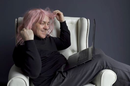 A man in a pink wig sits in a white chair and communicates via video link on a laptop. Side view