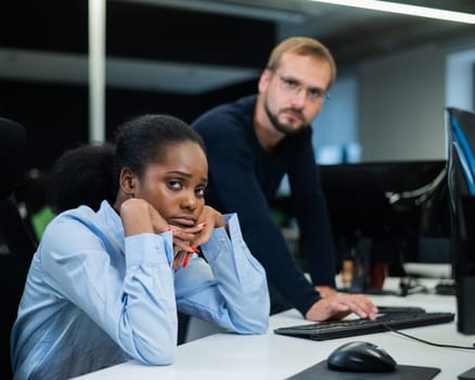 Colleagues look at the monitor and decide working moments. Caucasian man helps sad african woman solve computer problem