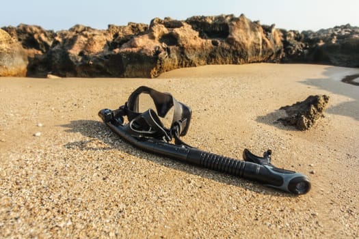 Black snorkel and diving mass on beach sand, small rocks in background. Shot through wet lens (drops make parts of picture blurry) to emphasise water.