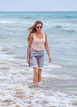 Sporty young woman in skirt, t shirts and sunglasses standing in shallow sea, small waves and overcast sky behind her.