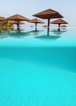 Wooden square sun parasols seen from calm infinity pool. Partially underwater photo, bottom part is blue tiles floor - space for text. Relax / vacation concept photo.