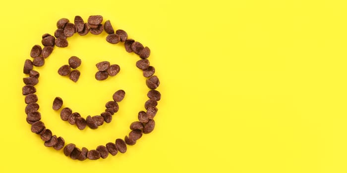 Tabletop view, smiley face made of brow chocolate corn flakes on yellow board. Space for text on right side.