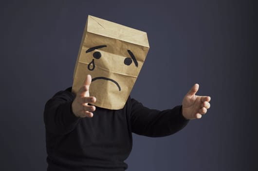 A sad man in a black turtleneck with a bag on his head, with a drawn crying emoticon, cries and asks for help stretching his hands forward. Emotions and gestures.