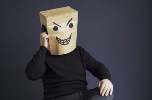 A man in a black turtleneck with a bag with an evil smiley on his head is talking on the phone while sitting on a chair. Emotions and anger