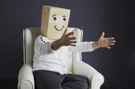 A man in a white shirt with a paper bag on his head, with a painted smiley, sits in an armchair, overwhelmed with feelings, stretches his arms for hugs.