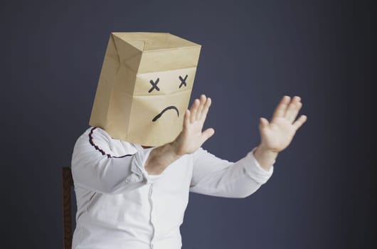 A sad man in a white shirt with a bag on his head, with a sad emoticon drawn, is afraid, defends himself by putting his hands forward. Emotions and gestures.