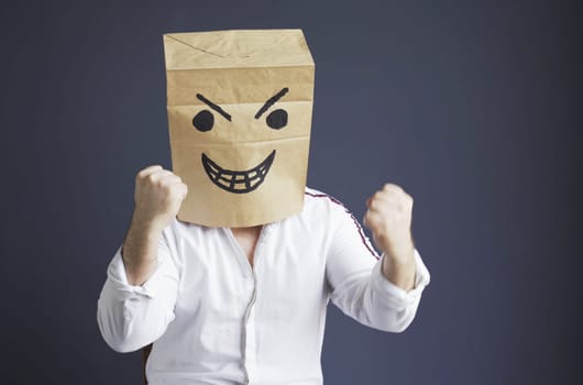A man in a white shirt with a paper bag with an angry emoticon on his head gesticulates aggressively with his hands. Emotions and anger