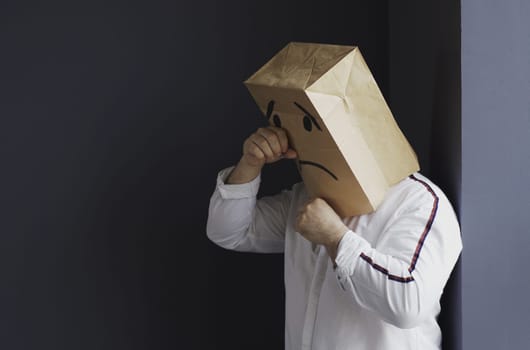 A sad man in a white shirt with a bag on his head, with a drawn crying emoticon, stands against the wall and cries. Emotions and gestures.